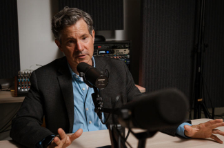 A person speaks into a podcast-style microphone inside an audio recording studio. Acoustic panels, recording equipment and a mixing console surround the workspace as the interview is being recorded.