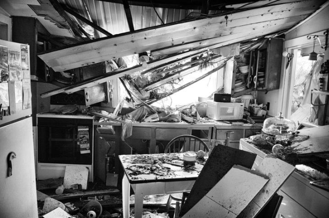 A kitchen severely damaged by a storm. The roof has collapsed inward, exposing beams and debris across counters, tables and appliances, illustrating the destructive impact of a natural disaster.